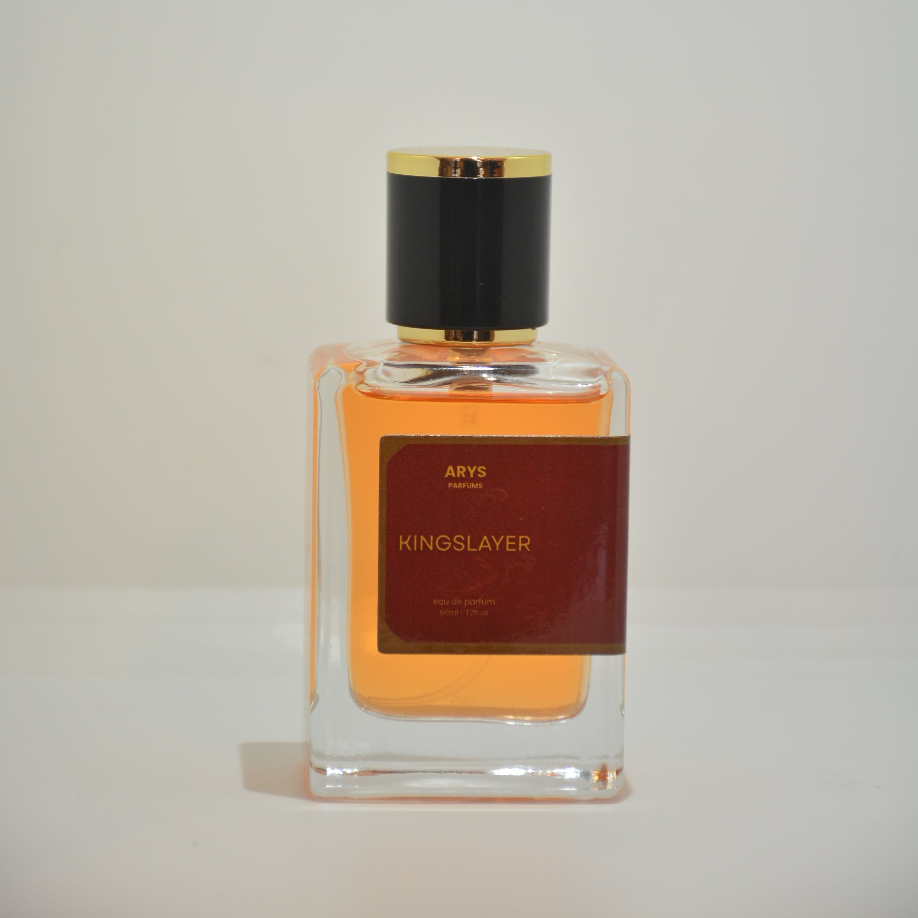 A bottle of Kingslayer perfume with amber liquid, a dark red label, and a black cap with gold trim, shown against a plain off-white background. Inspired by Bad Boy Elixir