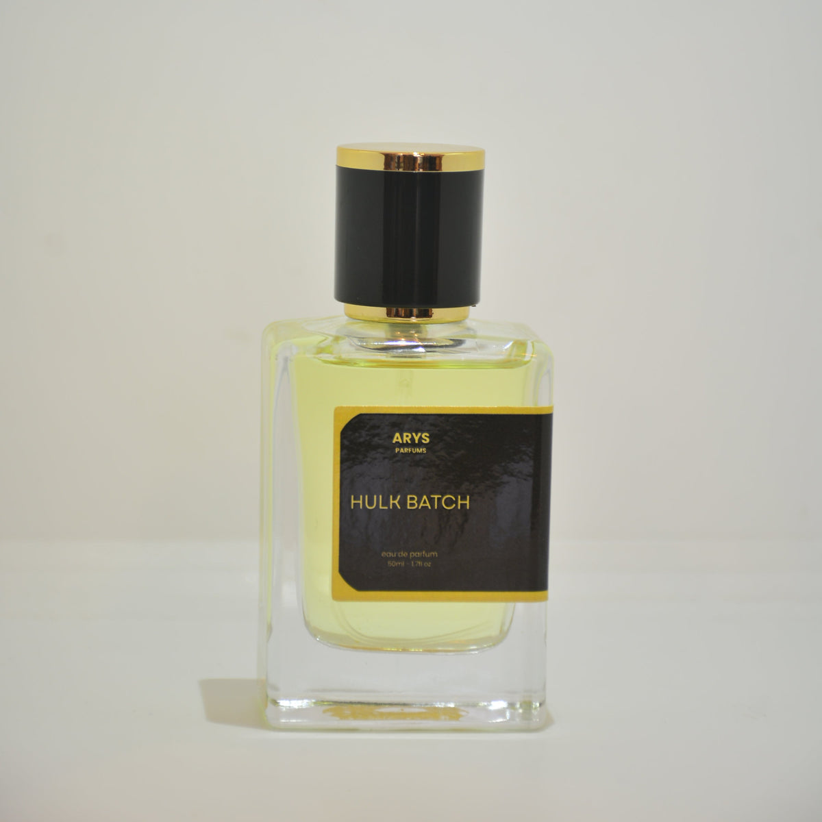 A clear rectangular glass perfume bottle filled with pale yellow liquid, featuring a black label with gold lettering and a black cylindrical cap with gold trim, photographed against a plain light background. This is Hulk Batch, inspired from the 2018 batch of Creed Aventus.