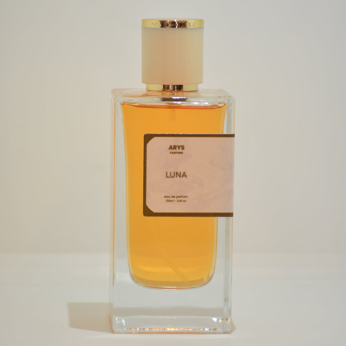 A rectangular clear glass perfume bottle filled with amber-yellow liquid, featuring a pale pink label with black text and a matching translucent cap, shown against a plain light background. This is Luna, inspired from Black Opium by YSL