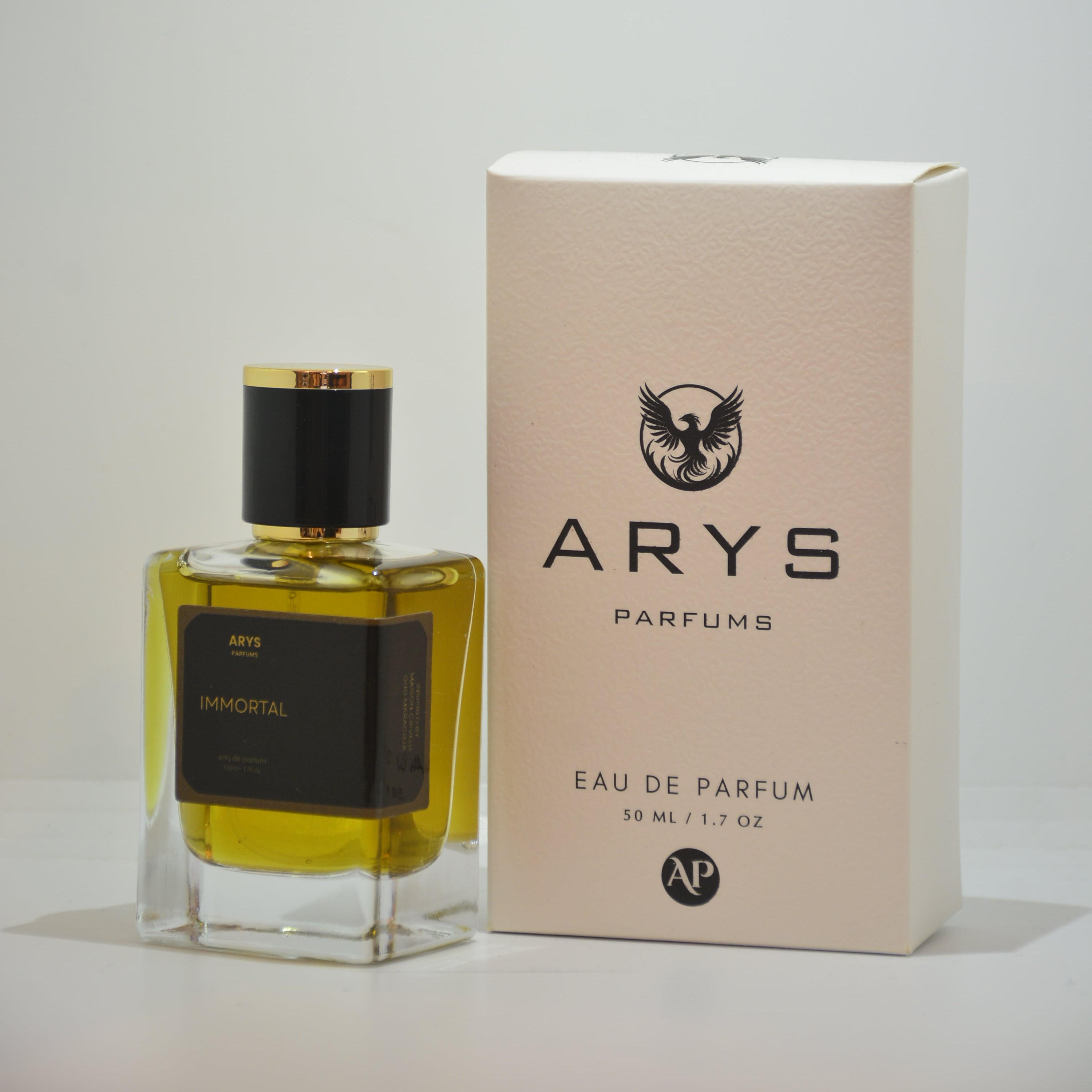 A bottle of Arys Parfums Immortal with a black cap and black label, next to its cream-colored packaging box, on a light gray background. This is Immortal, inspired from Oud Maracuja by Maison Crivelli.