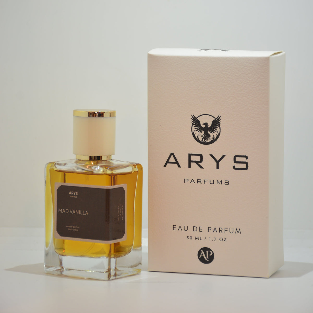 A bottle of Arys Parfums with amber liquid and a cream cap, shown next to its beige packaging box on a white background. It is Mad Vanilla, inspired from Vanilla 28 by Kayali.