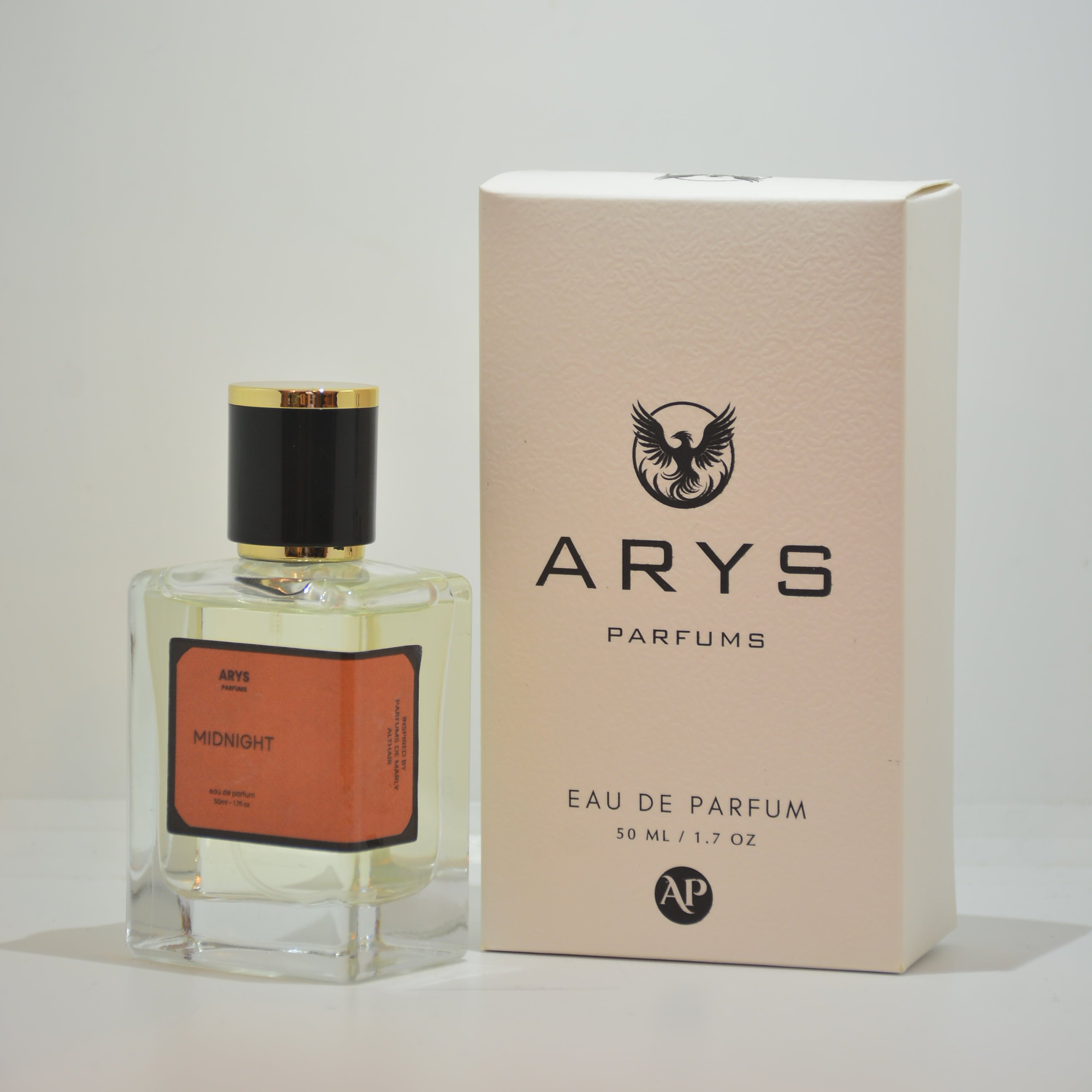 A bottle of Arys Parfum perfume with a clear glass body, yellow liquid, orange label, and black cap, shown next to its beige packaging box on a white background. This is Althair by Parfums De Marly.